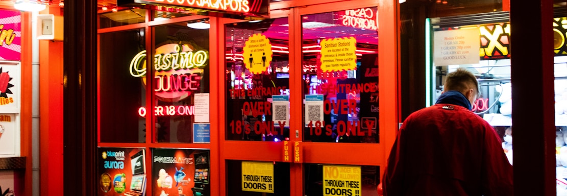 A brightly lit entrance to a casino with neon signs, including one advertising a £500 jackpot. The doors have clear signage indicating entry is restricted to those over 18. A person in a red jacket stands to the right, possibly interacting with a machine or display.